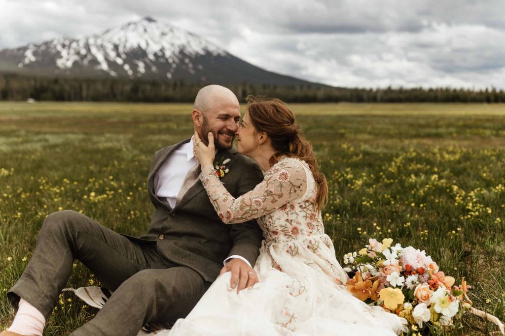 man and woman wearing wedding attire sitting in a field with a mountain in the background on their wedding day.