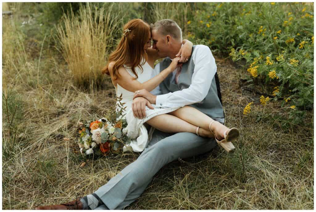 couple dressed in wedding attire sitting in a field and snuggling.