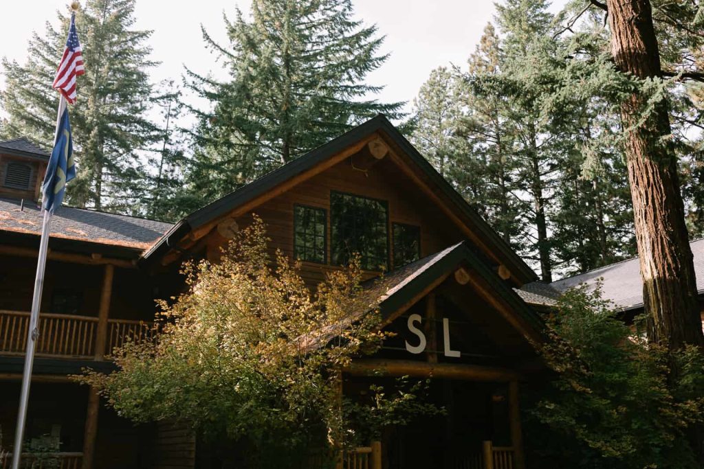 a rustic looking cabin with trees surrounding and an american flag to the left.