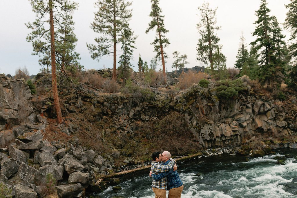 a couple standing above dillon falls embracing in a hug, with trees and water surrounding them.