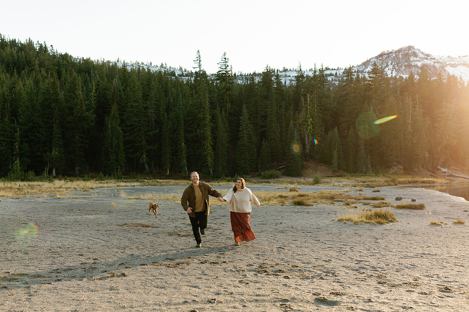 couple running hand in hand with dog behind them on a sandy beach with mountain in background